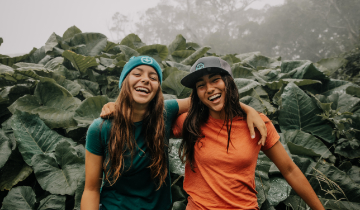 Two young women laughing with their arms around each other outside wearing Gather FCU hats